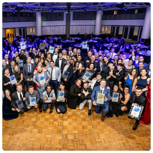 A large group of people are gathered in a formal event setting, posing for a group photo. Many individuals are holding plaques or awards, smiling and celebrating. The room is filled with round tables covered in white linens, suggesting a banquet or awards ceremony. The lighting is a vibrant blue, creating an energetic and celebratory atmosphere. Everyone is dressed in formal or semi-formal attire, and the mood appears to be festive and triumphant.