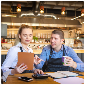 The image shows two people, likely restaurant or café staff, sitting at a table with various work materials. The woman is holding a tablet while the man points towards it, seemingly discussing or reviewing something. The setting appears to be a modern kitchen or café, with equipment visible in the background. Both are engaged in a professional conversation, with notebooks, a calculator, and a mobile phone on the table, suggesting they are working on business-related tasks.