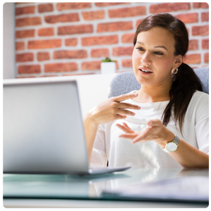 The image shows a woman sitting at a desk, engaged in a conversation or presentation via a video call on her laptop. She appears to be explaining or gesturing with her hands, and her expression is focused yet friendly. The setting is a modern home or office, with a brick wall in the background, creating a relaxed and professional atmosphere, suggesting an e-learning or virtual meeting environment.